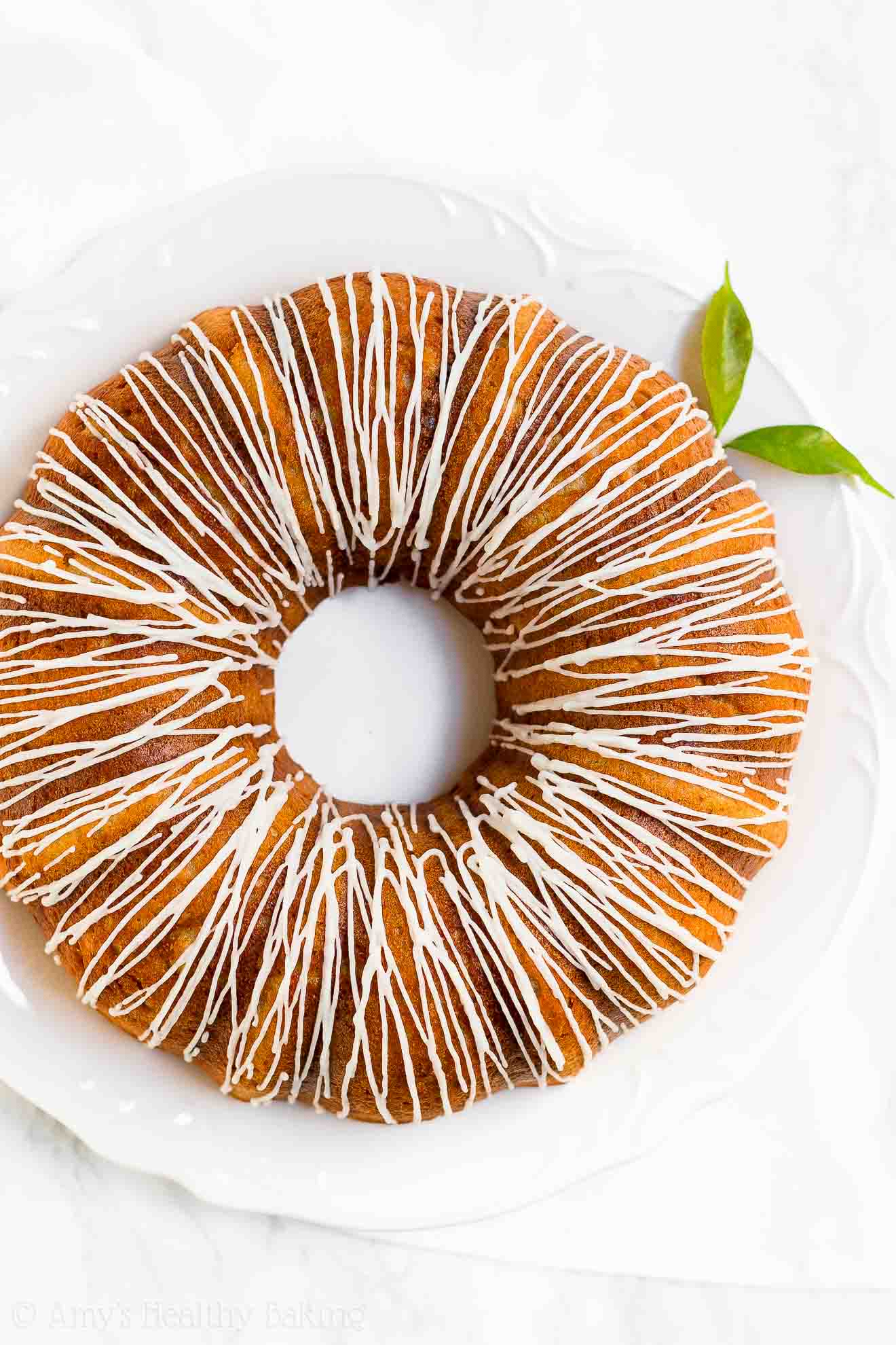 a freshly drizzled healthy lemon bundt cake on a large round white serving tray – easy lemon bundt cake recipe topped with glaze – moist, low calorie, low fat, sugar free, and clean eating with whole wheat and gluten free options – made with meyer lemons, greek yogurt, and no sugar