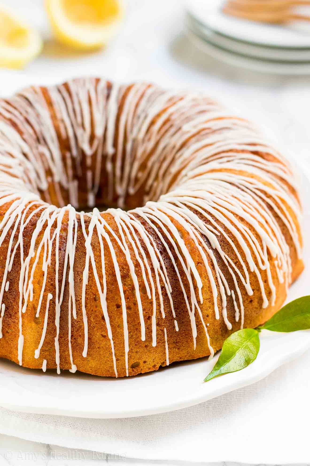 a healthy lemon bundt cake finished with thin lines of lemon drizzle on a large round white serving tray – easy lemon bundt cake recipe topped with glaze – moist, low calorie, low fat, sugar free, and clean eating with whole wheat and gluten free options – made with meyer lemons, greek yogurt, and no sugar