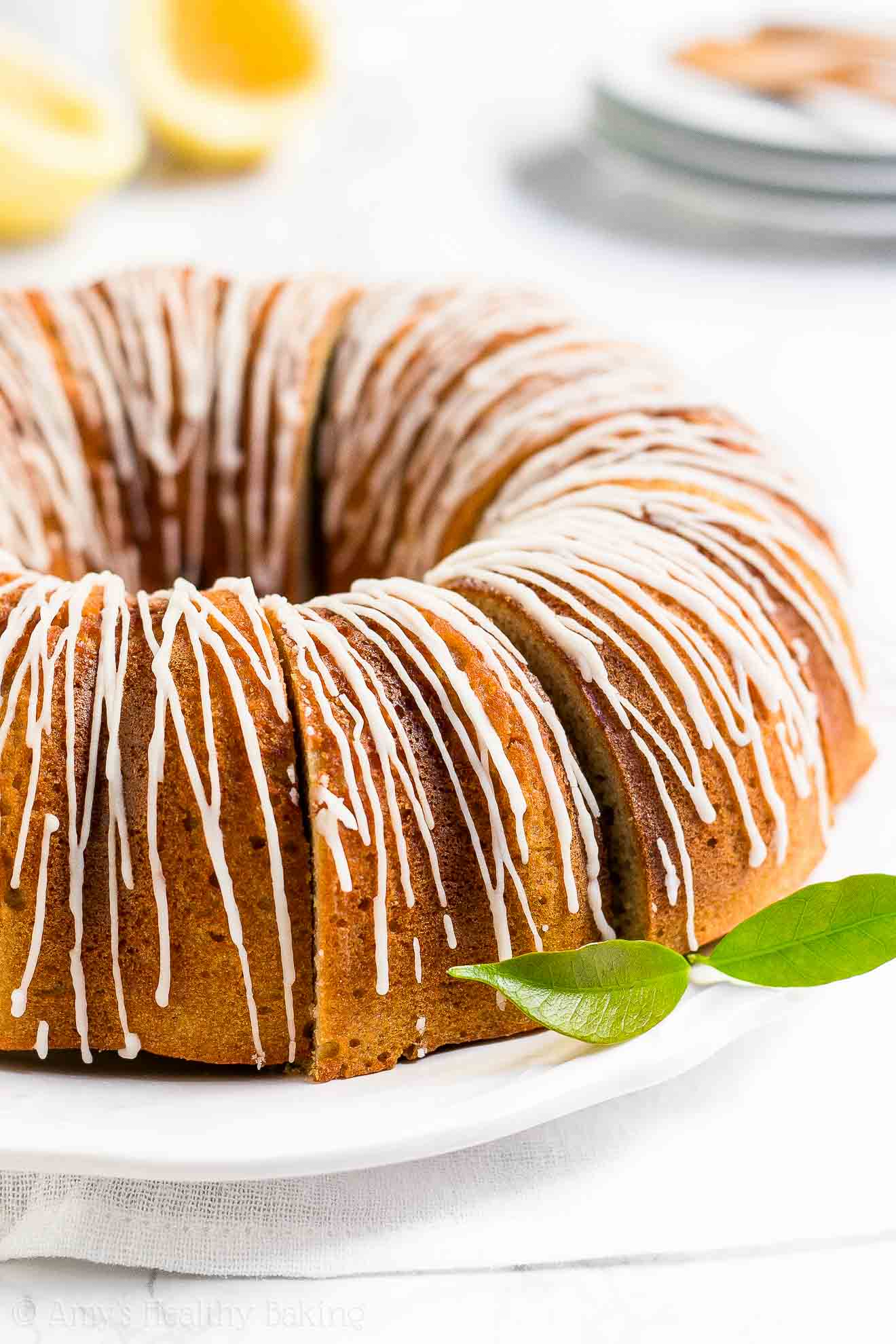 a partially sliced healthy lemon bundt cake on a large round white serving tray with small white plates and lemons in the background – easy lemon bundt cake recipe topped with glaze – moist, low calorie, low fat, sugar free, and clean eating with whole wheat and gluten free options – made with meyer lemons, greek yogurt, and no sugar