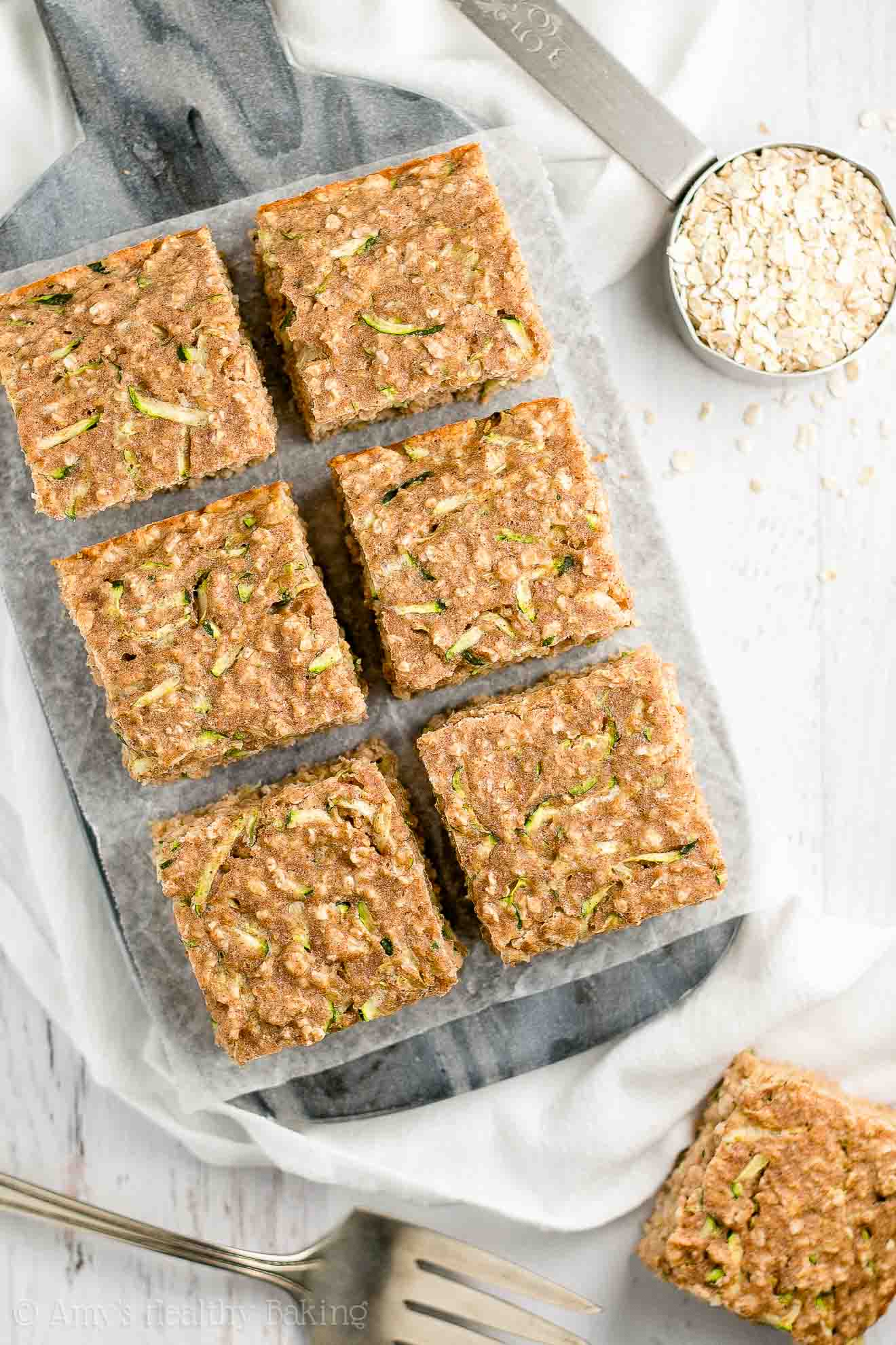 six square slices of healthy zucchini oatmeal snack cake on a dark marble serving tray surrounded by a measuring cup of oats, a serving utensil, and another square snack cake slice – easy recipe for a healthy zucchini cake full of oats and spices – low calorie, low fat, soft, moist, and clean eating with whole wheat and gluten free options – made with greek yogurt, pure maple syrup, and no refined sugar