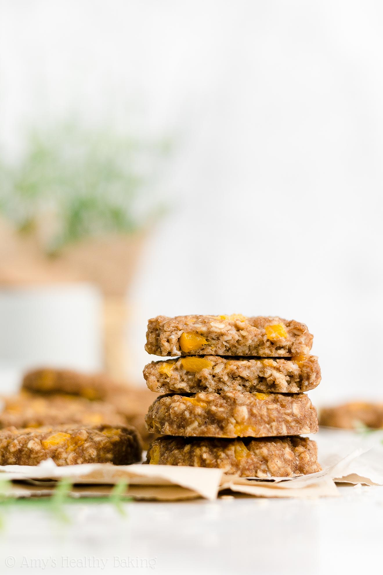 a stack of four of the best healthy peach oatmeal breakfast cookies, with the top two cookies broken in half to show the texture inside, and more cookies visible in the background – easy fruit and oat breakfast cookie recipe – soft, chewy, low calorie, low fat, dairy free, and clean eating with whole wheat and gluten free options – made with oats, spices, pure maple syrup, fresh peaches or canned peaches, and no refined sugar