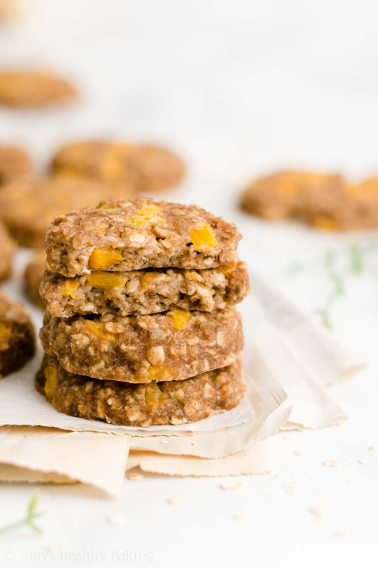 a stack of four of the best healthy peach oatmeal breakfast cookies resting on a folded newspaper, with the top two cookies broken in half to reveal the texture inside, and more cookies visible in the background – easy fruit and oat breakfast cookie recipe – soft, chewy, low calorie, low fat, dairy free, and clean eating with whole wheat and gluten free options – made with oats, spices, pure maple syrup, fresh peaches or canned peaches, and no refined sugar