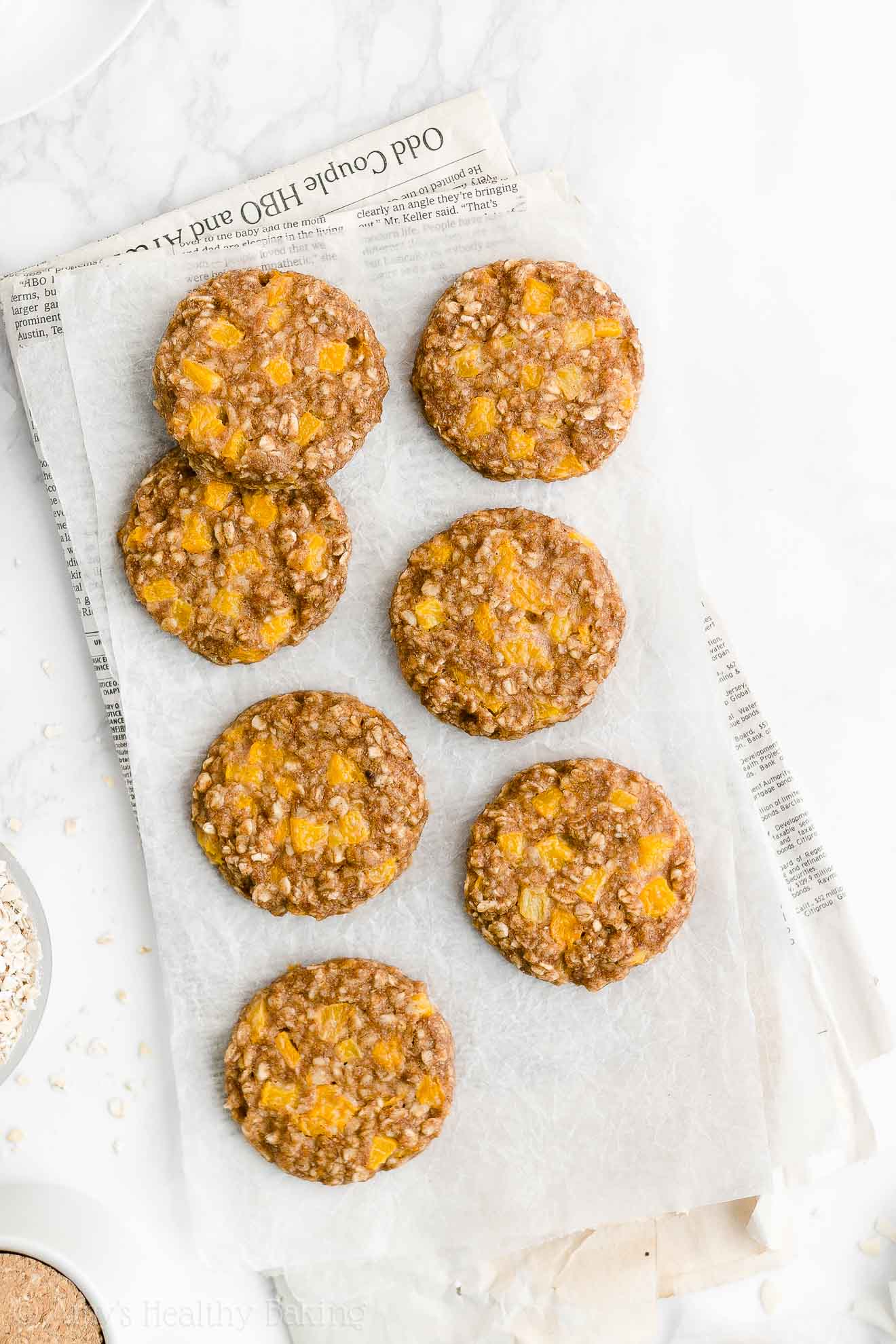 seven of the best healthy peach oatmeal breakfast cookies sitting on a rectangular piece of white parchment paper resting on top of a folded newspaper – easy fruit and oat breakfast cookie recipe – soft, chewy, low calorie, low fat, dairy free, and clean eating with whole wheat and gluten free options – made with oats, spices, pure maple syrup, fresh peaches or canned peaches, and no refined sugar