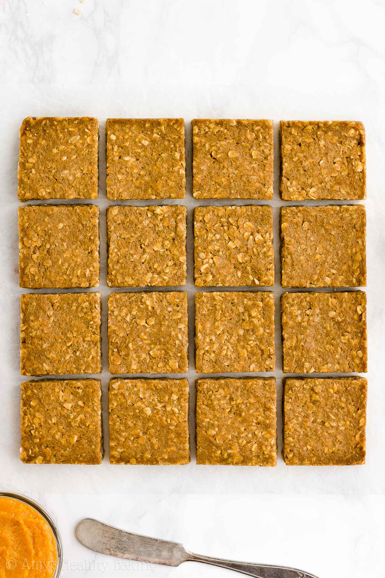 sixteen healthy pumpkin oatmeal bars neatly arranged into four tidy rows on a white marble countertop with a small bowl of pumpkin purée and a butter knife resting below them – easy oat bars recipe for breakfasts and snacks – vegan, gluten free, egg free, dairy free, and clean eating with whole wheat and gluten free options – made with oats, pumpkin, spices, and no refined sugar