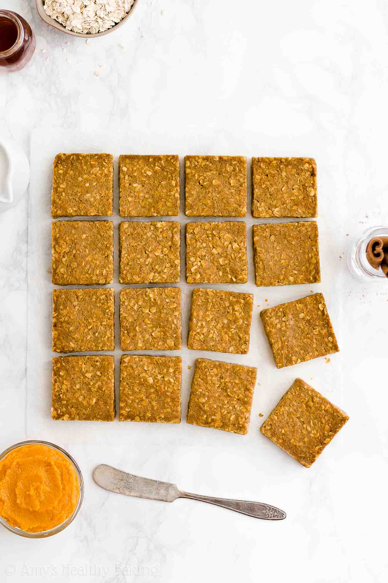 healthy pumpkin oatmeal bars on a white marble countertop and surrounded by a small bowl of oats, glass jar of maple syrup, jug of milk, bowl of pumpkin, butter knife, and cinnamon sticks – easy oat bars recipe for breakfasts and snacks – vegan, gluten free, egg free, dairy free, and clean eating with whole wheat and gluten free options – made with oats, pumpkin, spices, and no refined sugar