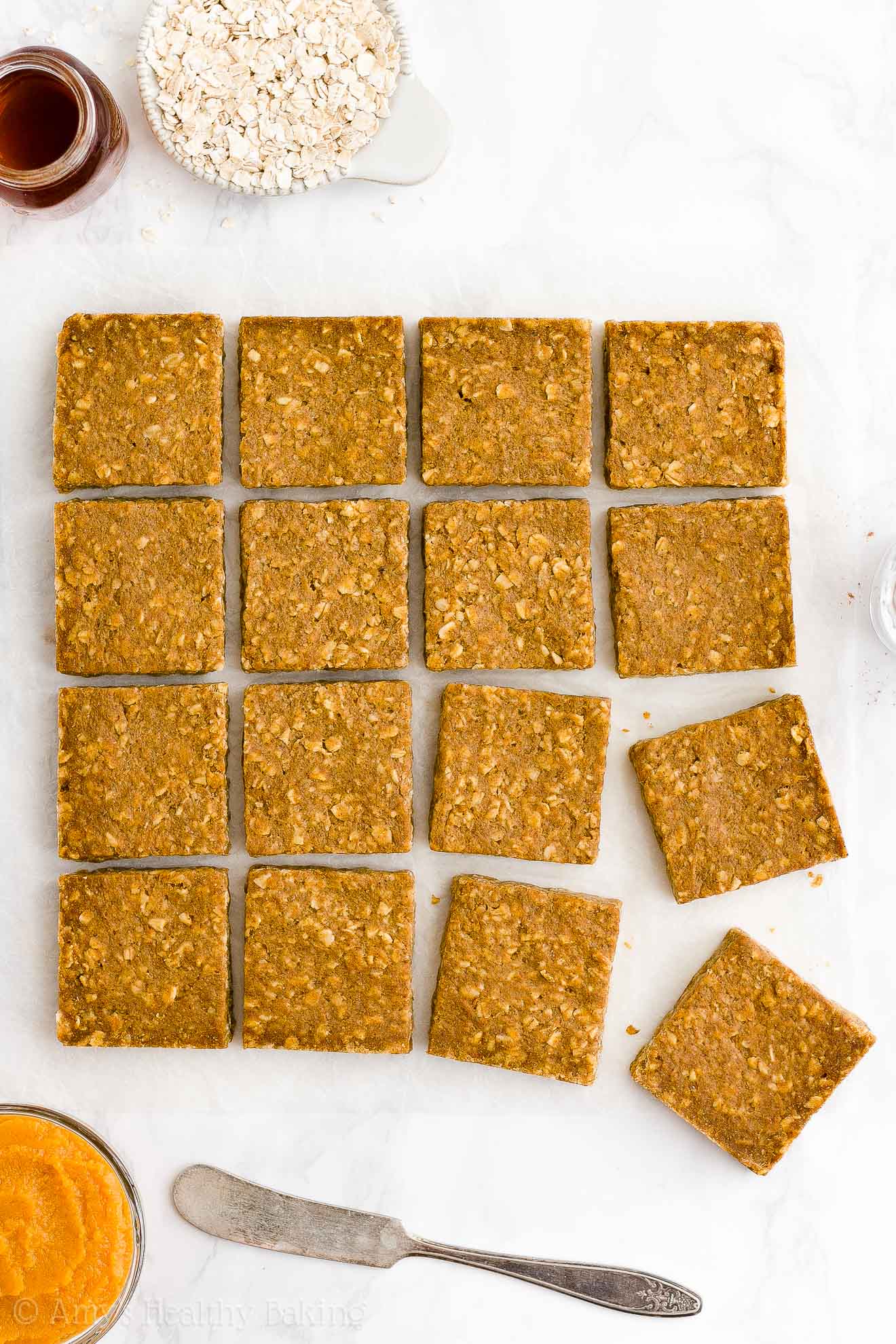 overhead view of healthy pumpkin oatmeal bars on a white marble countertop and surrounded by small bowls of oats, pumpkin, maple syrup, and a butter knife – easy oat bars recipe for breakfasts and snacks – vegan, gluten free, egg free, dairy free, and clean eating with whole wheat and gluten free options – made with oats, pumpkin, spices, and no refined sugar