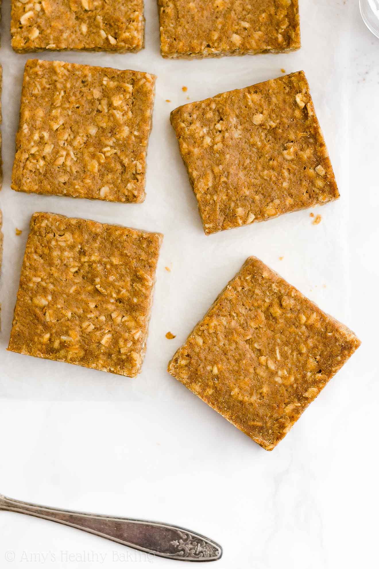 overhead view of healthy pumpkin oatmeal bars on white parchment paper, with the handle of a butter knife visible in the bottom left corner – easy oat bars recipe for breakfasts and snacks – vegan, gluten free, egg free, dairy free, and clean eating with whole wheat and gluten free options – made with oats, pumpkin, spices, and no refined sugar