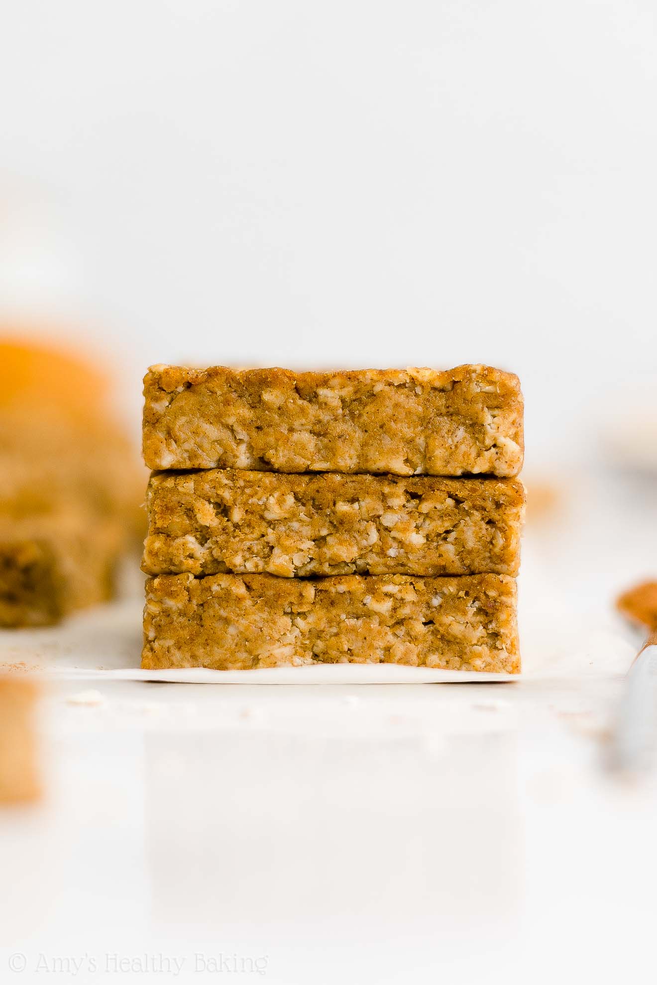stack of three healthy pumpkin oatmeal bars on white parchment paper with more bars in the background – easy oat bars recipe for breakfasts and snacks – vegan, gluten free, egg free, dairy free, and clean eating with whole wheat and gluten free options – made with oats, pumpkin, spices, and no refined sugar