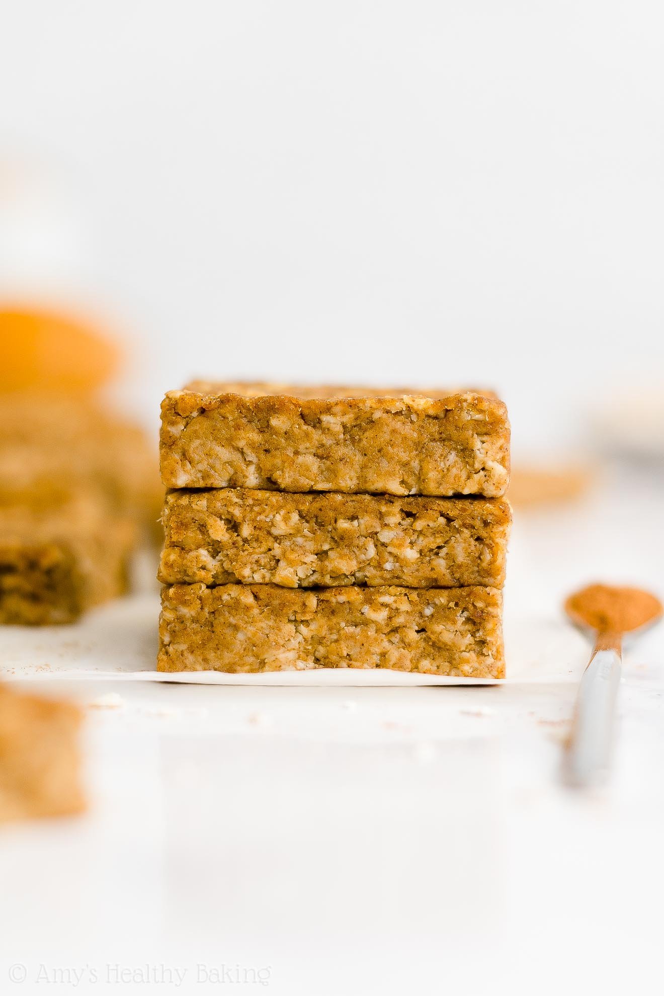 stack of three healthy pumpkin oatmeal bars on white parchment paper alongside a small spoon holding cinnamon with more bars and a bowl of pumpkin visible in the background – easy oat bars recipe for breakfasts and snacks – vegan, gluten free, egg free, dairy free, and clean eating with whole wheat and gluten free options – made with oats, pumpkin, spices, and no refined sugar