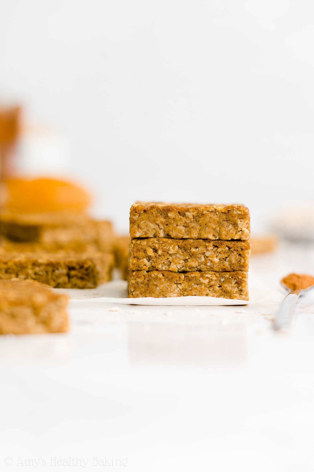 stack of three healthy pumpkin oatmeal bars on white parchment paper alongside a small spoonful of cinnamon with more bars and ingredients visible in the background – easy oat bars recipe for breakfasts and snacks – vegan, gluten free, egg free, dairy free, and clean eating with whole wheat and gluten free options – made with oats, pumpkin, spices, and no refined sugar