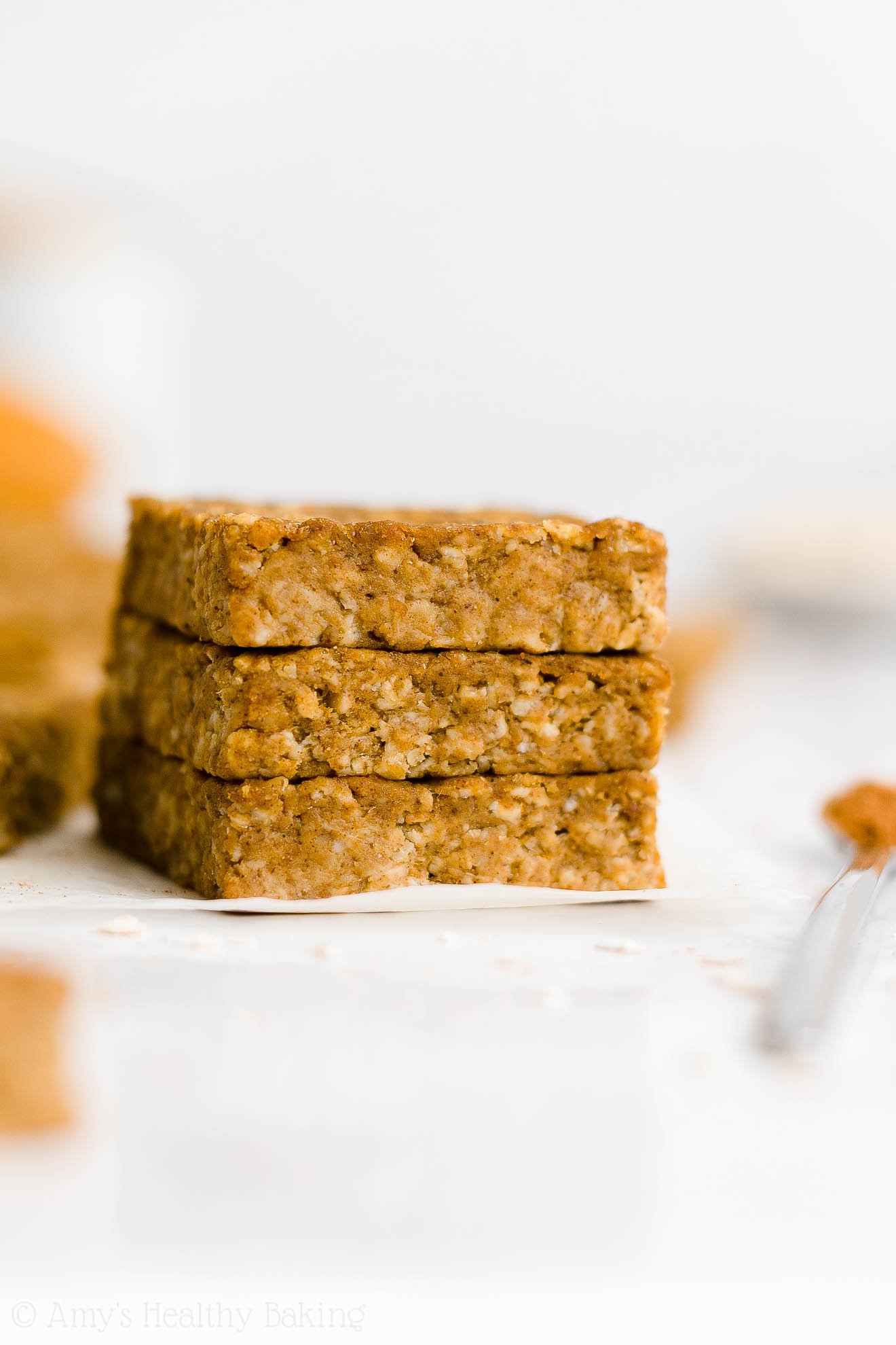 stack of three healthy pumpkin oatmeal bars on white parchment paper turned slightly to the right, with a small spoon holding cinnamon to their right – easy oat bars recipe for breakfasts and snacks – vegan, gluten free, egg free, dairy free, and clean eating with whole wheat and gluten free options – made with oats, pumpkin, spices, and no refined sugar