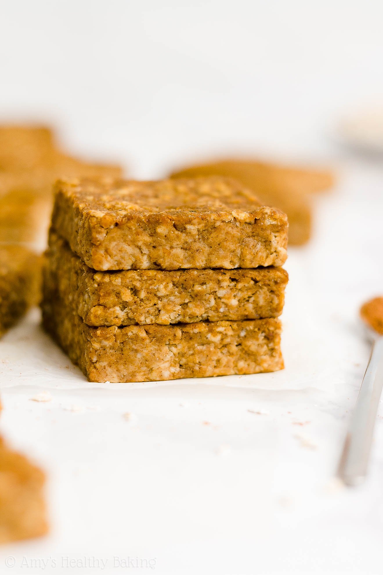 stack of three healthy pumpkin oatmeal bars on white parchment paper alongside a small spoonful of cinnamon with more bars visible behind them – easy oat bars recipe for breakfasts and snacks – vegan, gluten free, egg free, dairy free, and clean eating with whole wheat and gluten free options – made with oats, pumpkin, spices, and no refined sugar