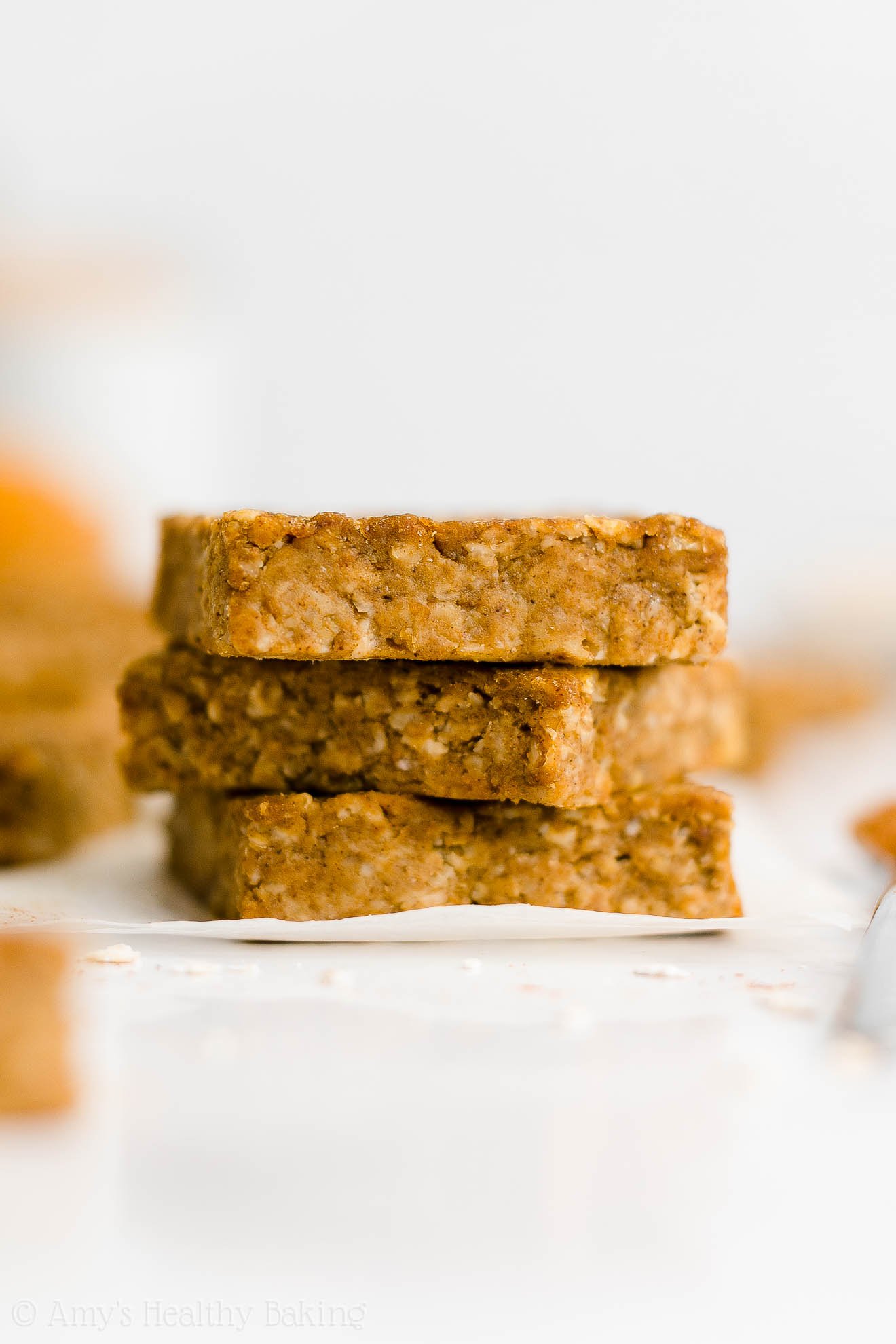 stack of three healthy pumpkin oatmeal bars on white parchment paper turned at slightly different angles with more bars visible in the background – easy oat bars recipe for breakfasts and snacks – vegan, gluten free, egg free, dairy free, and clean eating with whole wheat and gluten free options – made with oats, pumpkin, spices, and no refined sugar