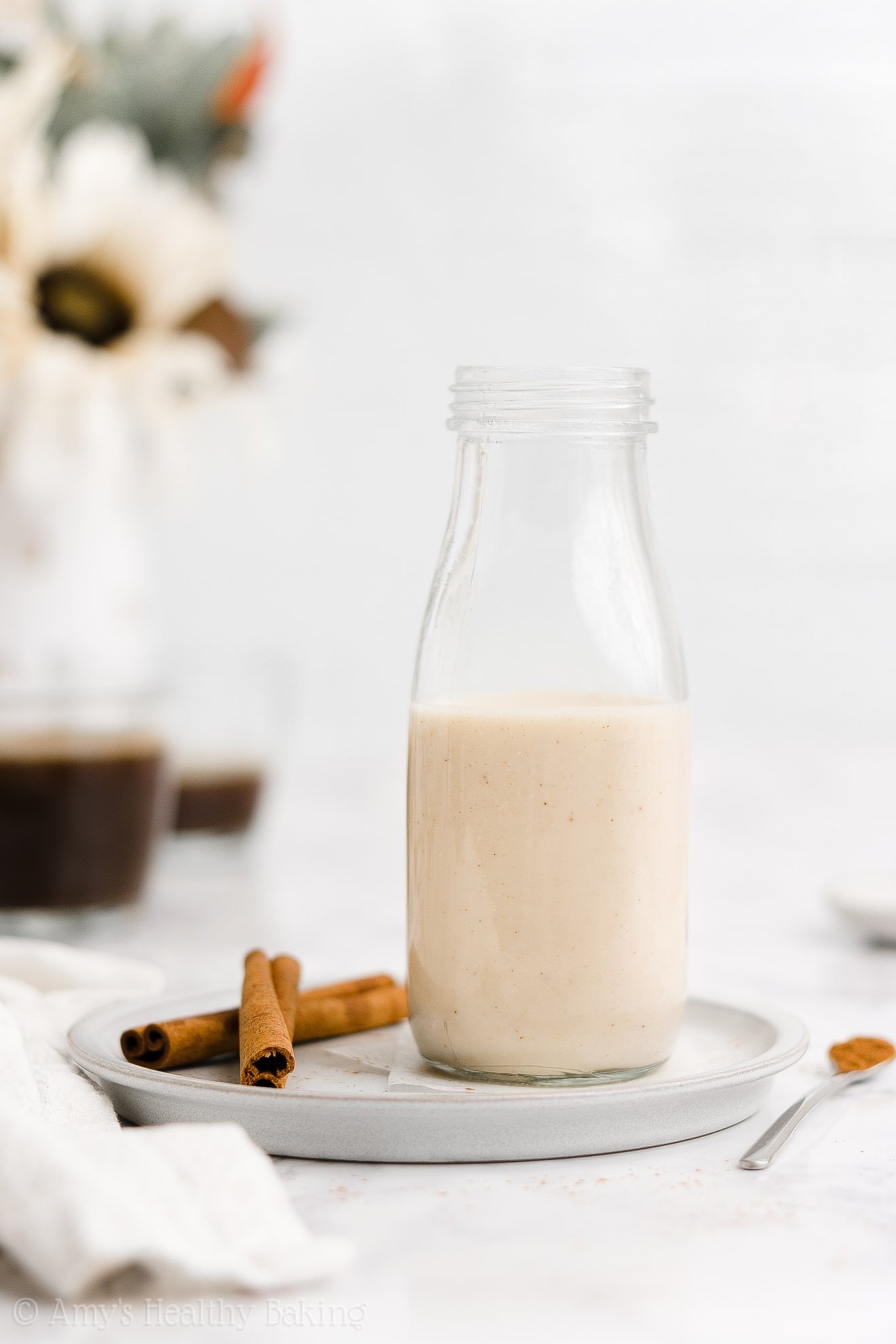 glass milk bottle holding sugar free gingerbread coffee creamer while sitting on a small gray plate beside two cinnamon sticks with two mugs of coffee in the background – healthy homemade gingerbread coffee creamer recipe – dairy free, vegan, low calorie, low fat, and clean eating – made with unsweetened cashew milk, spices, stevia, no sugar, and no molasses