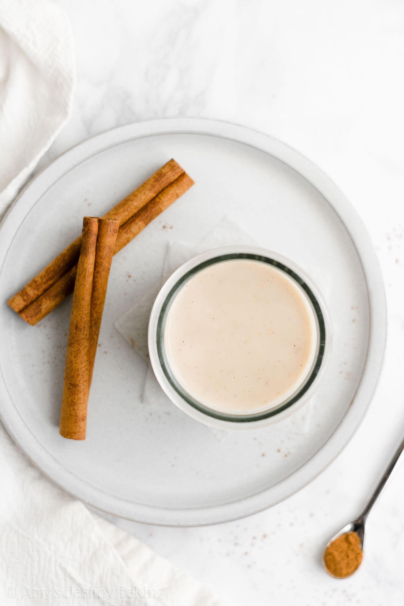 overhead view of a gray plate topped with a glass bottle holding sugar free gingerbread coffee creamer with two cinnamon sticks beside it – healthy homemade gingerbread coffee creamer recipe – dairy free, vegan, low calorie, low fat, and clean eating – made with unsweetened cashew milk, spices, stevia, no sugar, and no molasses