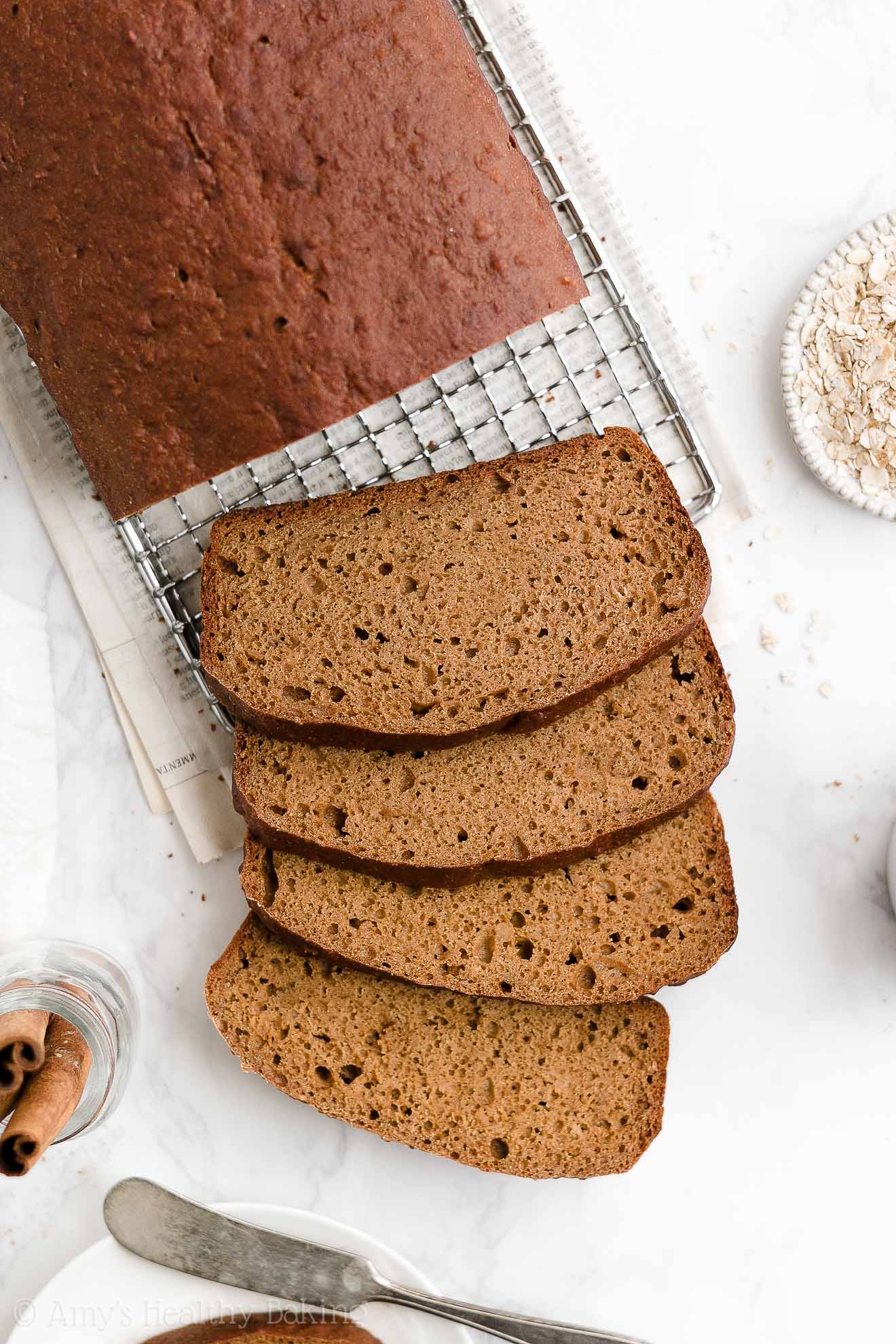 a healthy oatmeal gingerbread loaf on a wire cooling rack with four slices cut and falling down, also surrounded by a small bowl of oats, a jar of cinnamon sticks, and another slice on a white plate paired with a butter knife – easy gingerbread quick bread recipe – soft, moist, low fat, low calorie, and clean eating with whole wheat and gluten free options – made with oats, Greek yogurt, molasses, and no refined sugar