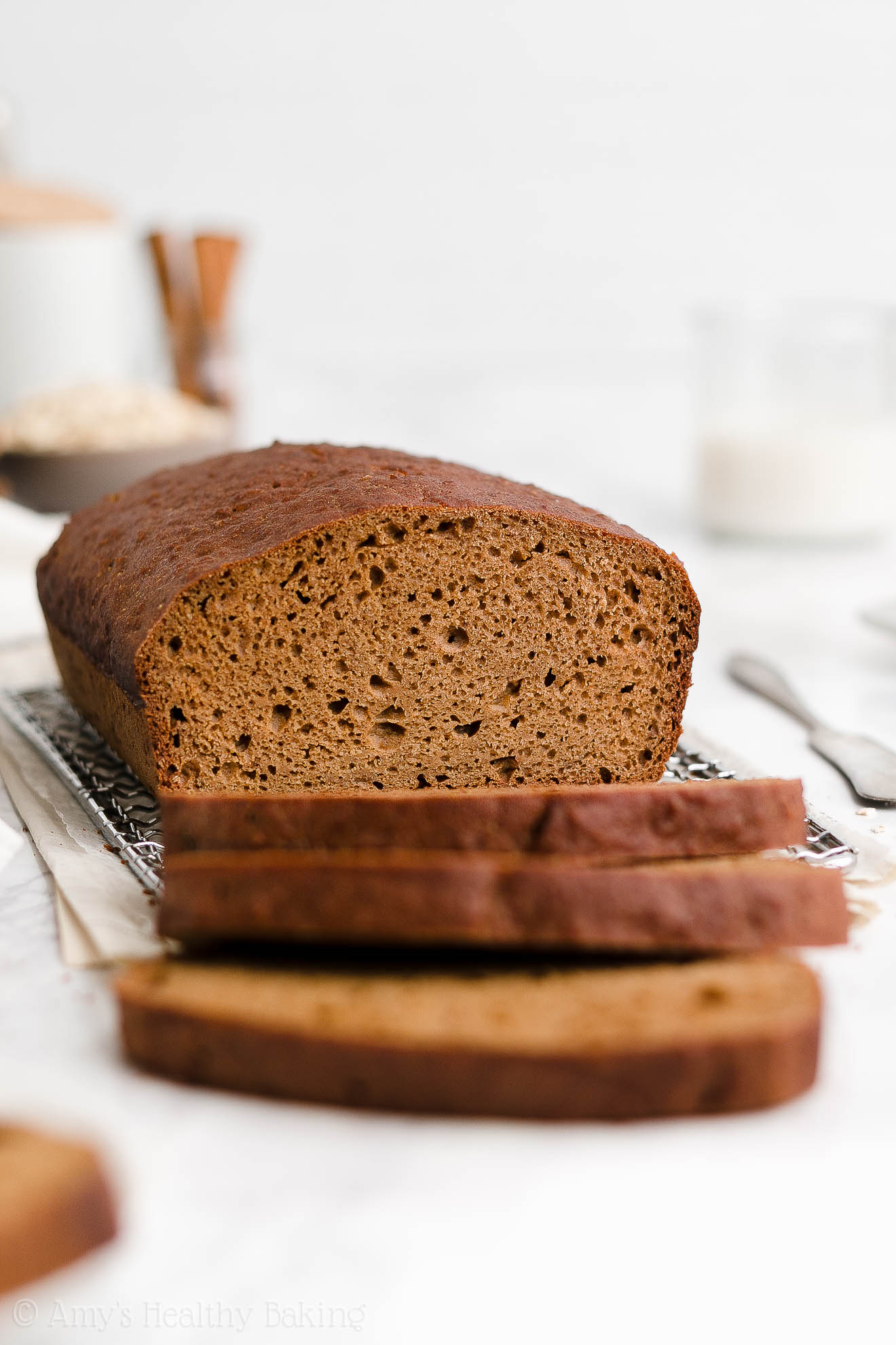 a healthy oatmeal gingerbread loaf sitting on a wire cooling rack with three slices freshly cut and falling over, plus a glass of milk and some ingredients visible in the background – easy gingerbread quick bread recipe – soft, moist, low fat, low calorie, and clean eating with whole wheat and gluten free options – made with oats, Greek yogurt, molasses, and no refined sugar