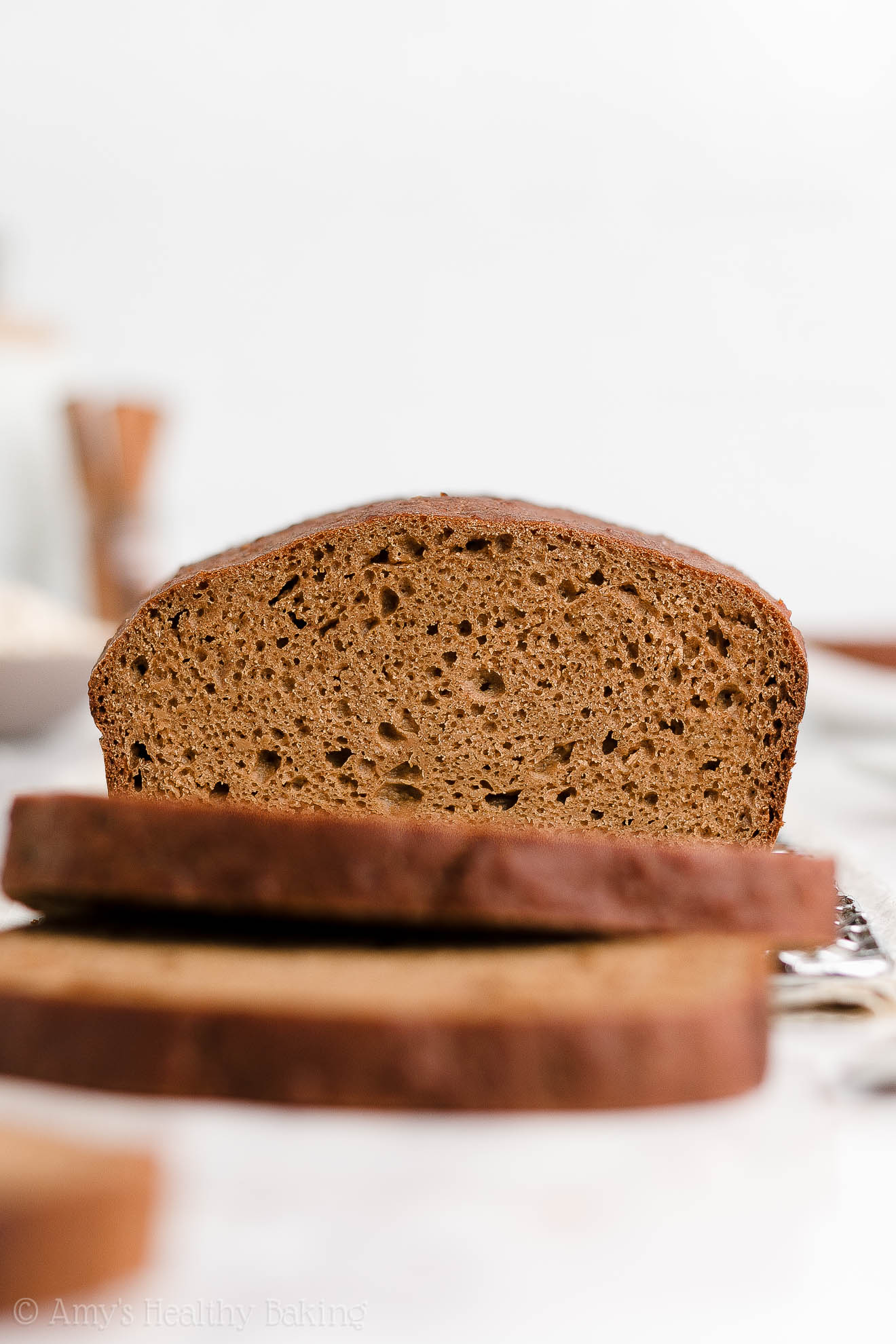 a partially sliced healthy oatmeal gingerbread loaf sitting on a wire cooling rack with two slices in the foreground directly in front of it – easy gingerbread quick bread recipe – soft, moist, low fat, low calorie, and clean eating with whole wheat and gluten free options – made with oats, Greek yogurt, molasses, and no refined sugar