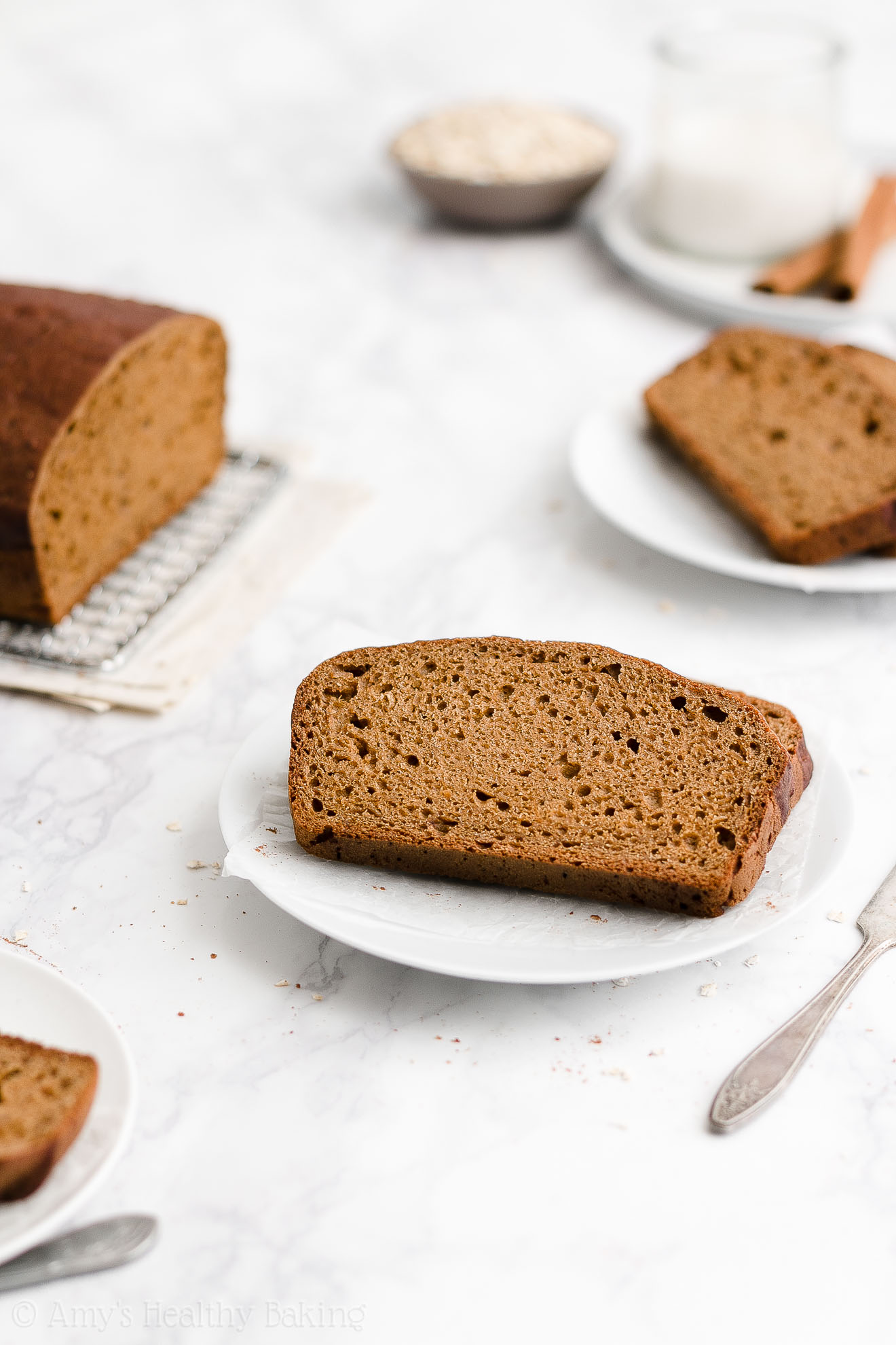 two slices of healthy oatmeal gingerbread loaf on a small white plate with another plate of slices, a glass of milk, and a bowl of oats in the background – easy gingerbread quick bread recipe – soft, moist, low fat, low calorie, and clean eating with whole wheat and gluten free options – made with oats, Greek yogurt, molasses, and no refined sugar
