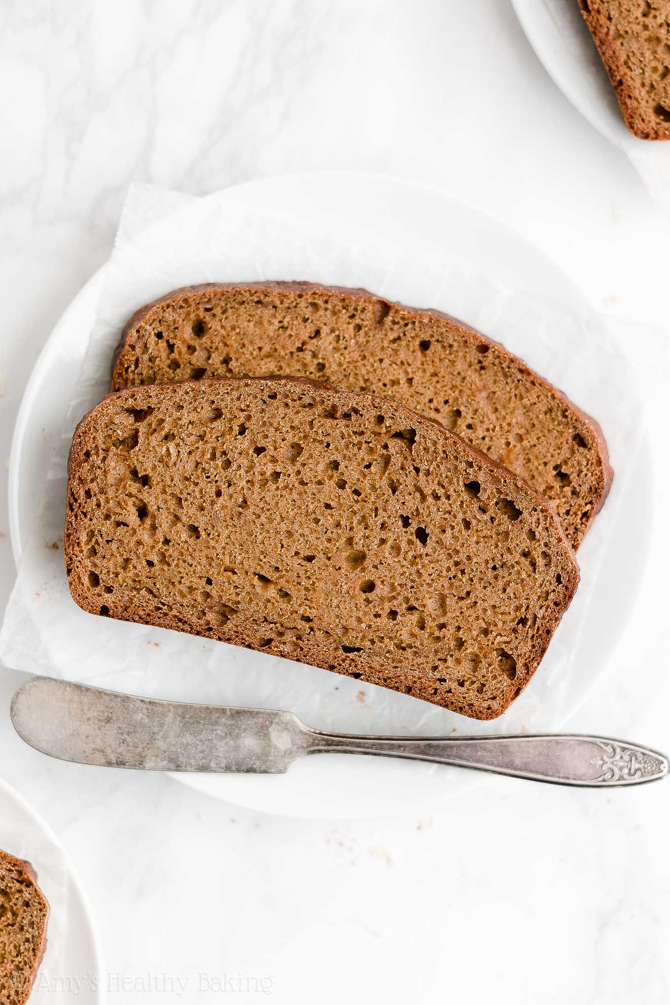 two slices of healthy oatmeal gingerbread loaf sitting on a small white plate with a butter knife resting beneath them – easy gingerbread quick bread recipe – soft, moist, low fat, low calorie, and clean eating with whole wheat and gluten free options – made with oats, Greek yogurt, molasses, and no refined sugar