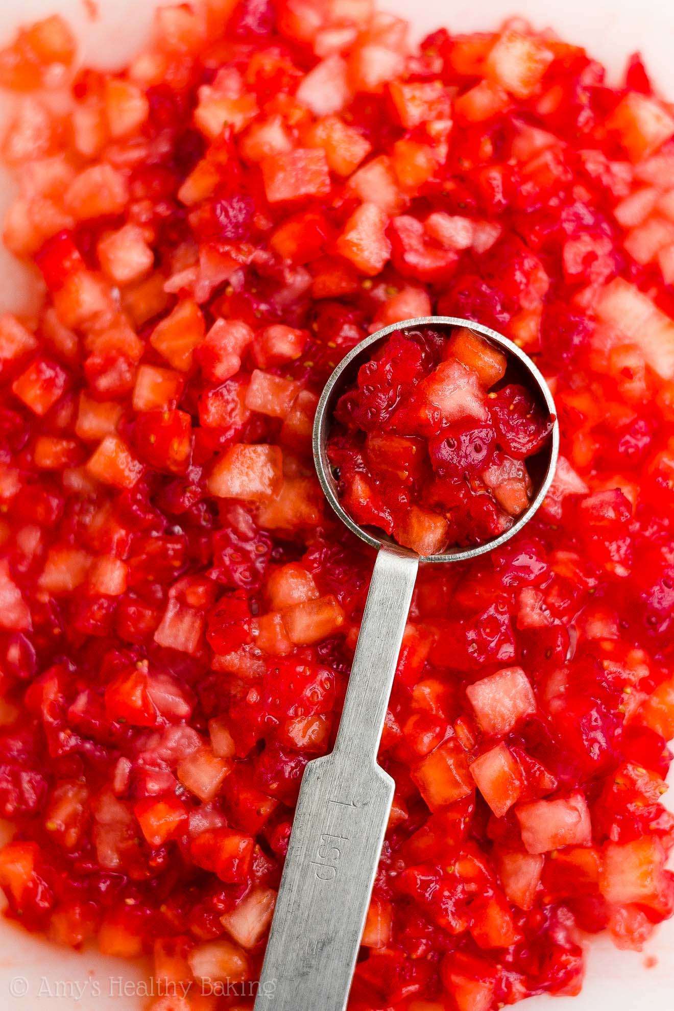 a 1-teaspoon measuring spoon holding diced strawberries and resting on top of a pile of diced strawberries, about to be turned into healthy strawberry oatmeal breakfast cookies – easy strawberry breakfast cookies recipe – soft, chewy, low calorie, low fat, and clean eating with whole wheat, gluten free, dairy free, and nut free options – made with oats, fresh strawberries, pure maple syrup, and no refined sugar