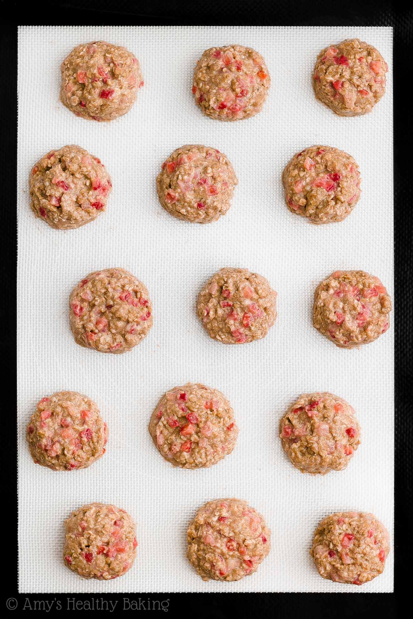 15 rounded mounds of raw healthy strawberry oatmeal breakfast cookie dough on a black and white silicone baking mat – easy strawberry breakfast cookies recipe – soft, chewy, low calorie, low fat, and clean eating with whole wheat, gluten free, dairy free, and nut free options – made with oats, fresh strawberries, pure maple syrup, and no refined sugar