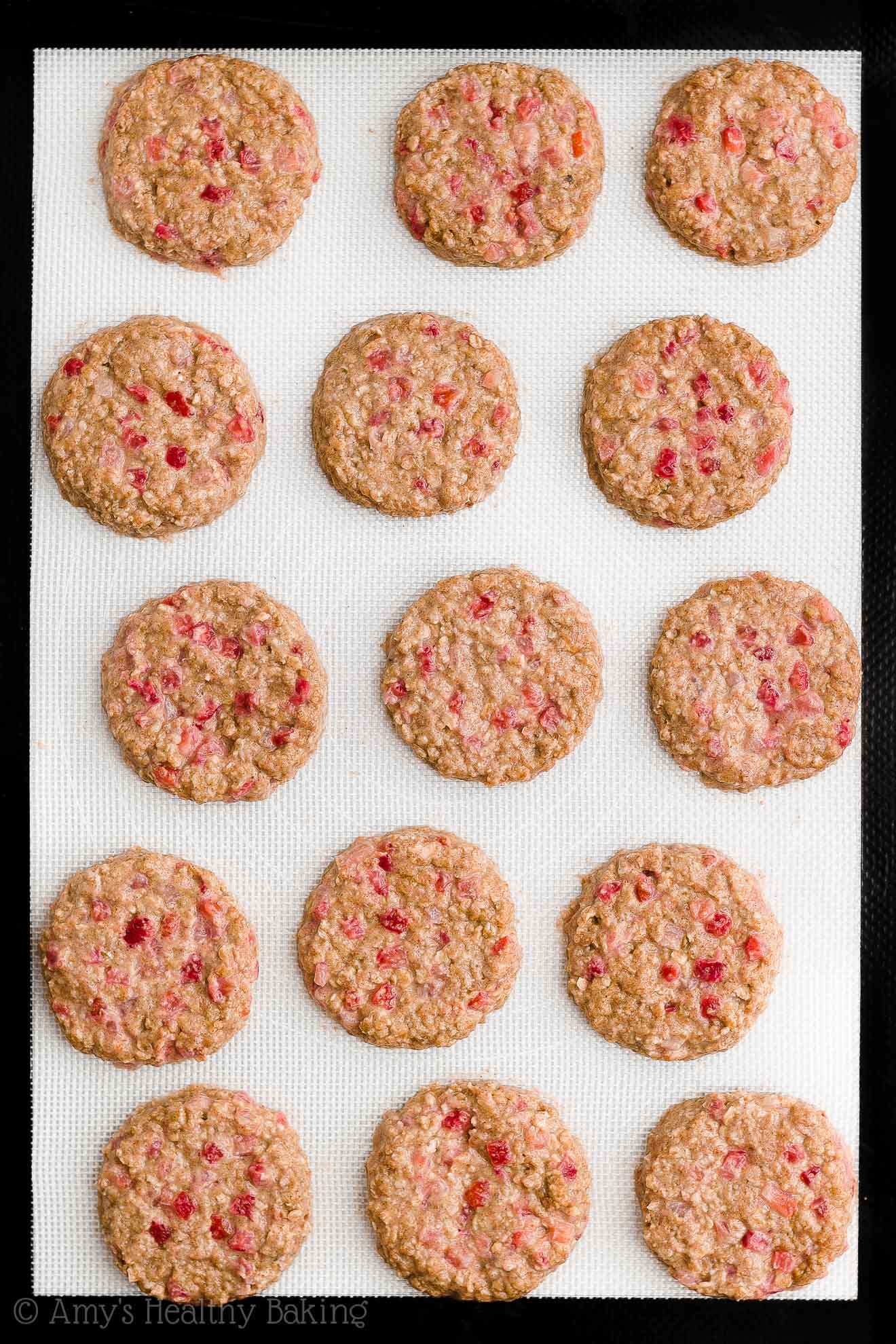 15 flattened discs of healthy strawberry oatmeal breakfast cookie dough on a black and white silicone baking mat – easy strawberry breakfast cookies recipe – soft, chewy, low calorie, low fat, and clean eating with whole wheat, gluten free, dairy free, and nut free options – made with oats, fresh strawberries, pure maple syrup, and no refined sugar