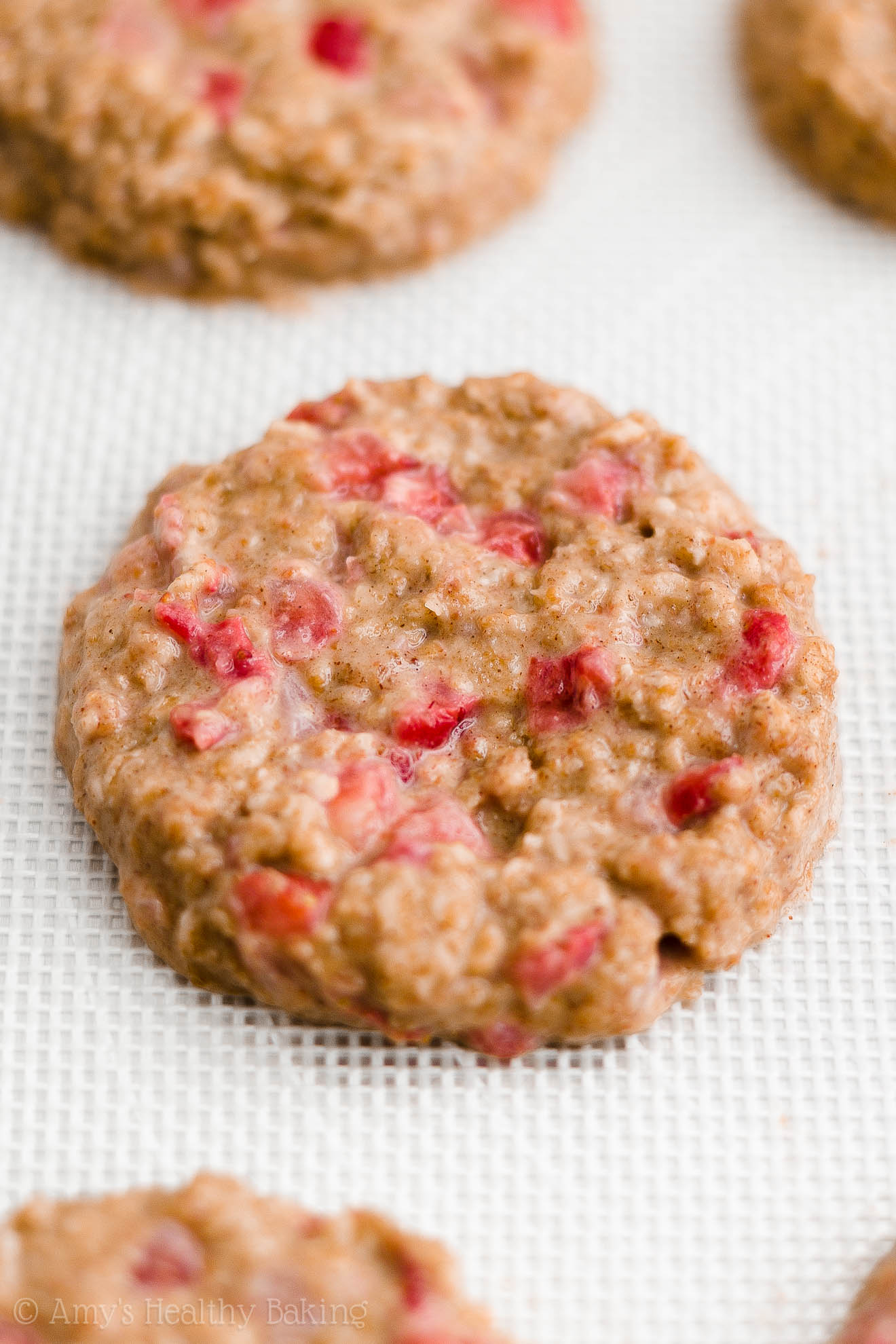 close-up of flattened discs of healthy strawberry oatmeal breakfast cookie dough on a white silicone baking mat – easy strawberry breakfast cookies recipe – soft, chewy, low calorie, low fat, and clean eating with whole wheat, gluten free, dairy free, and nut free options – made with oats, fresh strawberries, pure maple syrup, and no refined sugar