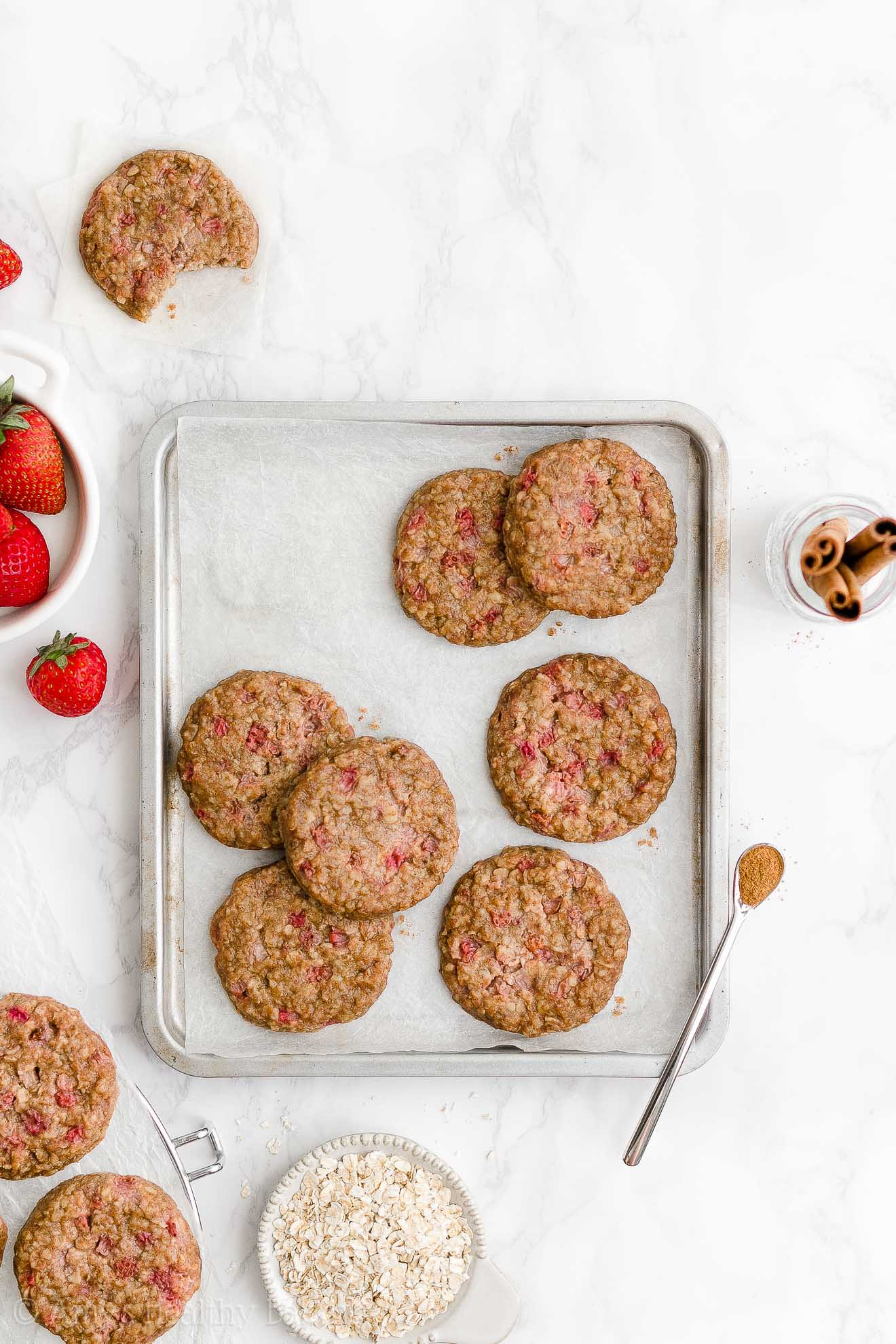 healthy strawberry oatmeal breakfast cookies arranged on a small rimmed baking sheet lined with white parchment paper and surrounded by a bowl of fresh strawberries, a bowl of oats, a jar of cinnamon sticks, and a few more cookies – easy strawberry breakfast cookies recipe – soft, chewy, low calorie, low fat, and clean eating with whole wheat, gluten free, dairy free, and nut free options – made with oats, fresh strawberries, pure maple syrup, and no refined sugar