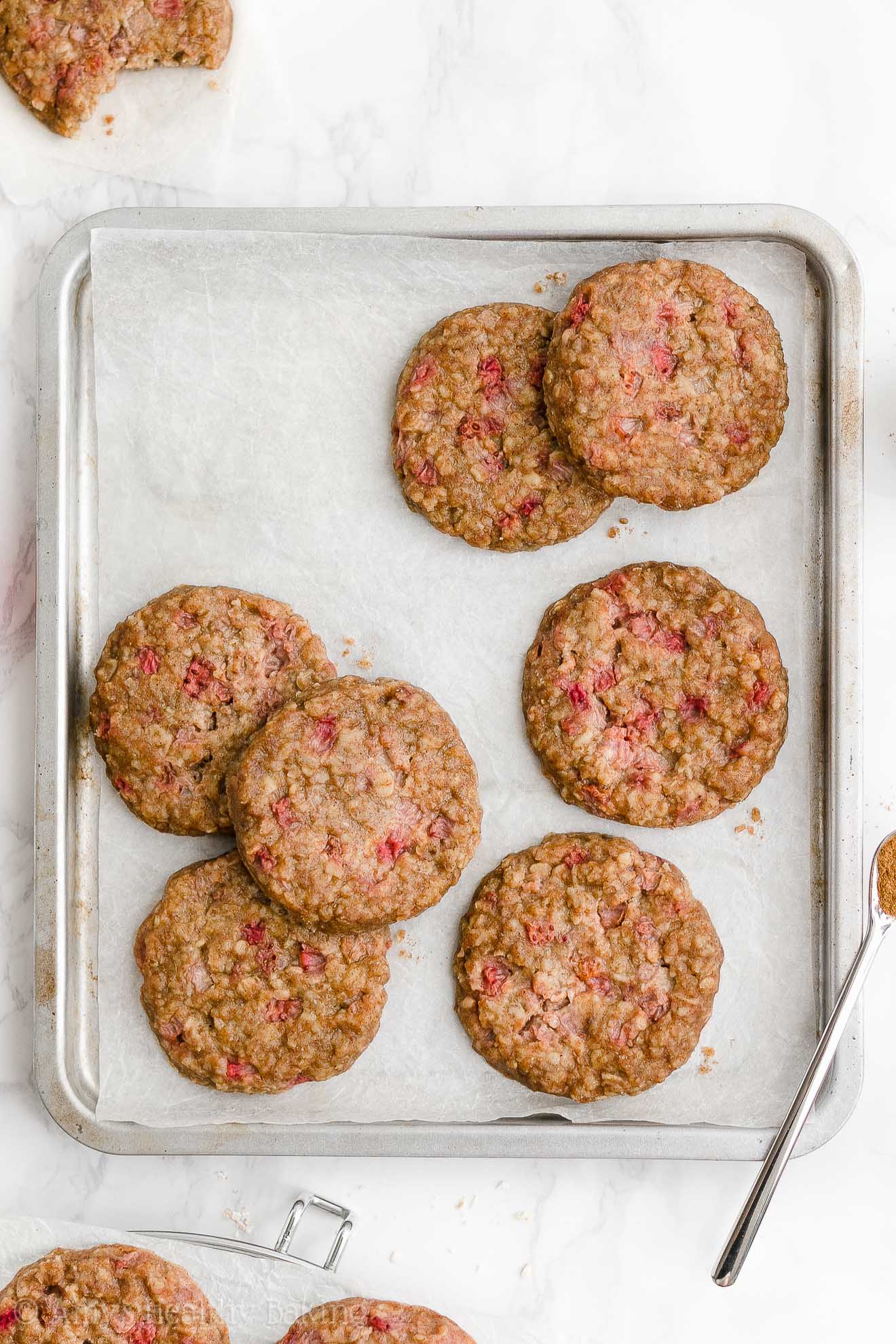 healthy strawberry oatmeal breakfast cookies arranged on a small rimmed baking sheet lined with white parchment paper surrounded by a few more cookies on parchment paper and a wire cooling rack – easy strawberry breakfast cookies recipe – soft, chewy, low calorie, low fat, and clean eating with whole wheat, gluten free, dairy free, and nut free options – made with oats, fresh strawberries, pure maple syrup, and no refined sugar