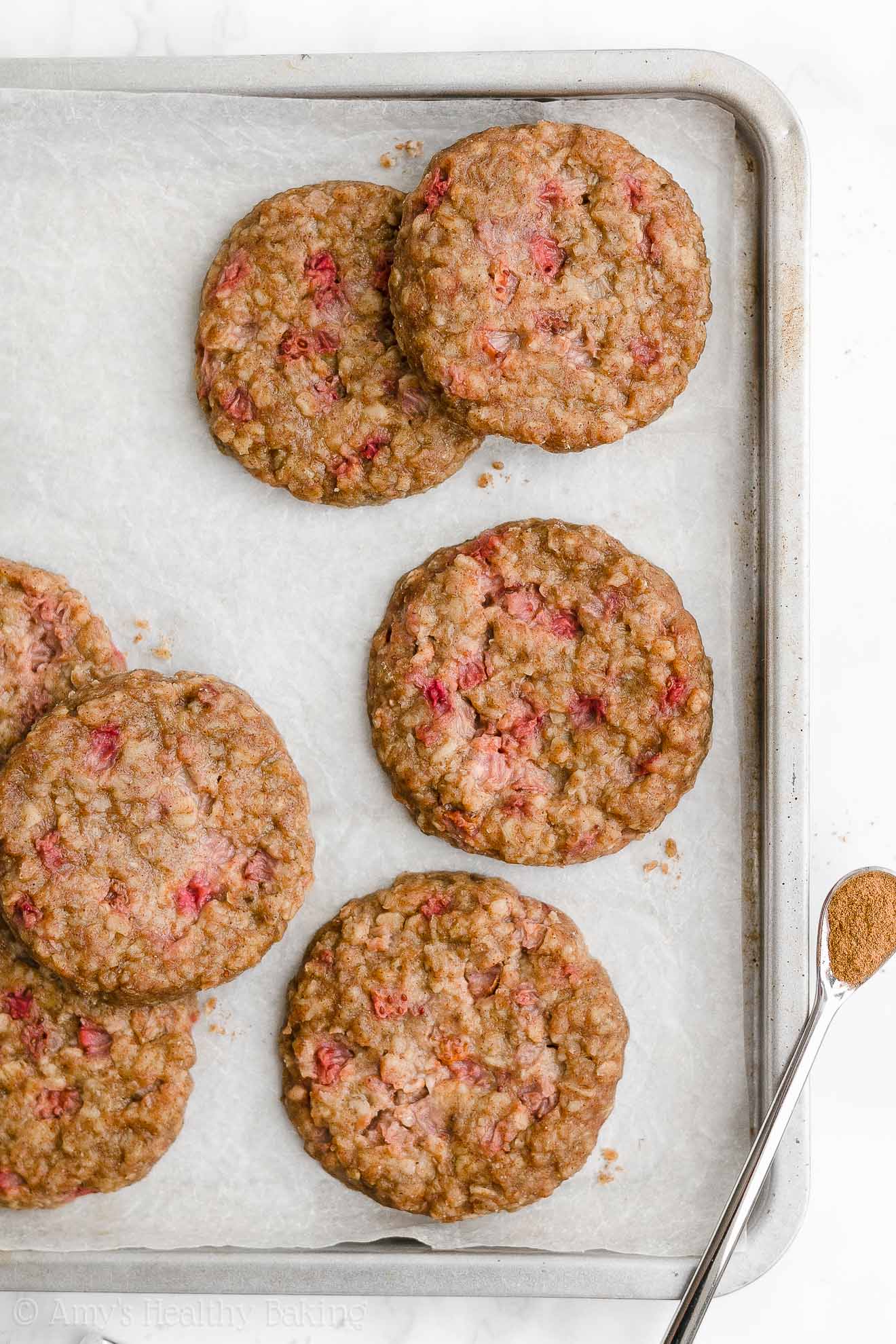 healthy strawberry oatmeal breakfast cookies arranged inside of a rimmed baking sheet lined with white parchment paper, with a small spoonful of cinnamon resting on the bottom right corner – easy strawberry breakfast cookies recipe – soft, chewy, low calorie, low fat, and clean eating with whole wheat, gluten free, dairy free, and nut free options – made with oats, fresh strawberries, pure maple syrup, and no refined sugar