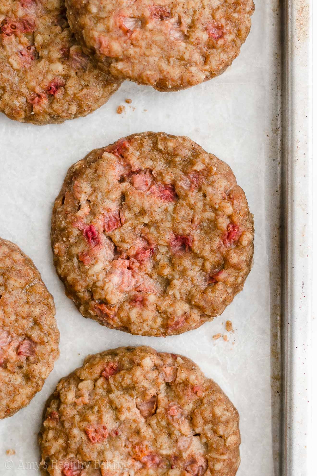 healthy strawberry oatmeal breakfast cookies on white parchment paper inside of a light metal rimmed baking sheet – easy strawberry breakfast cookies recipe – soft, chewy, low calorie, low fat, and clean eating with whole wheat, gluten free, dairy free, and nut free options – made with oats, fresh strawberries, pure maple syrup, and no refined sugar