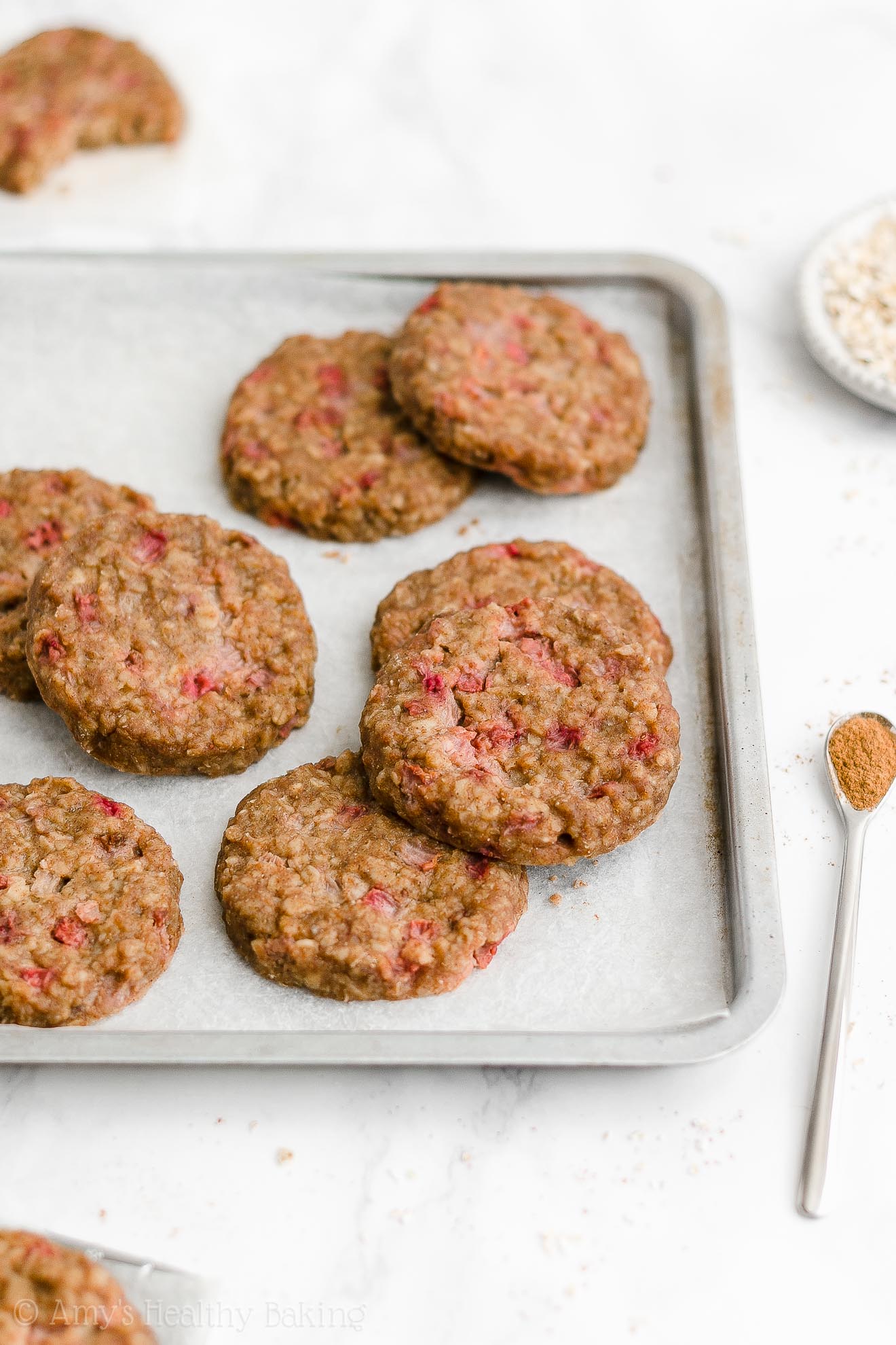 healthy strawberry oatmeal breakfast cookies arranged inside of a rimmed baking sheet lined with white parchment paper, with a small bowl of oats and another cookie visible in the background – easy strawberry breakfast cookies recipe – soft, chewy, low calorie, low fat, and clean eating with whole wheat, gluten free, dairy free, and nut free options – made with oats, fresh strawberries, pure maple syrup, and no refined sugar