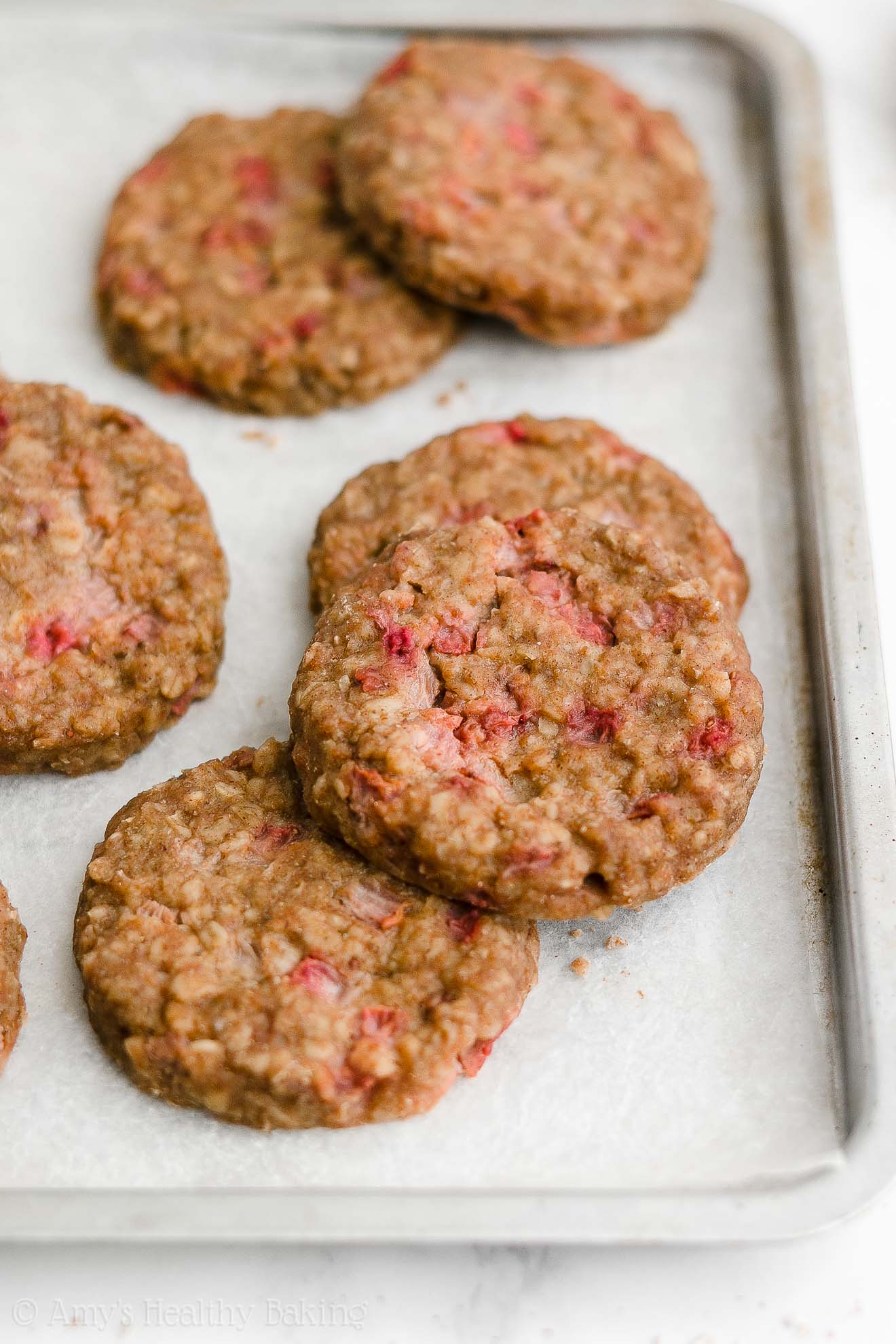 healthy strawberry oatmeal breakfast cookies arranged inside of a rimmed baking sheet lined with white parchment paper – easy strawberry breakfast cookies recipe – soft, chewy, low calorie, low fat, and clean eating with whole wheat, gluten free, dairy free, and nut free options – made with oats, fresh strawberries, pure maple syrup, and no refined sugar