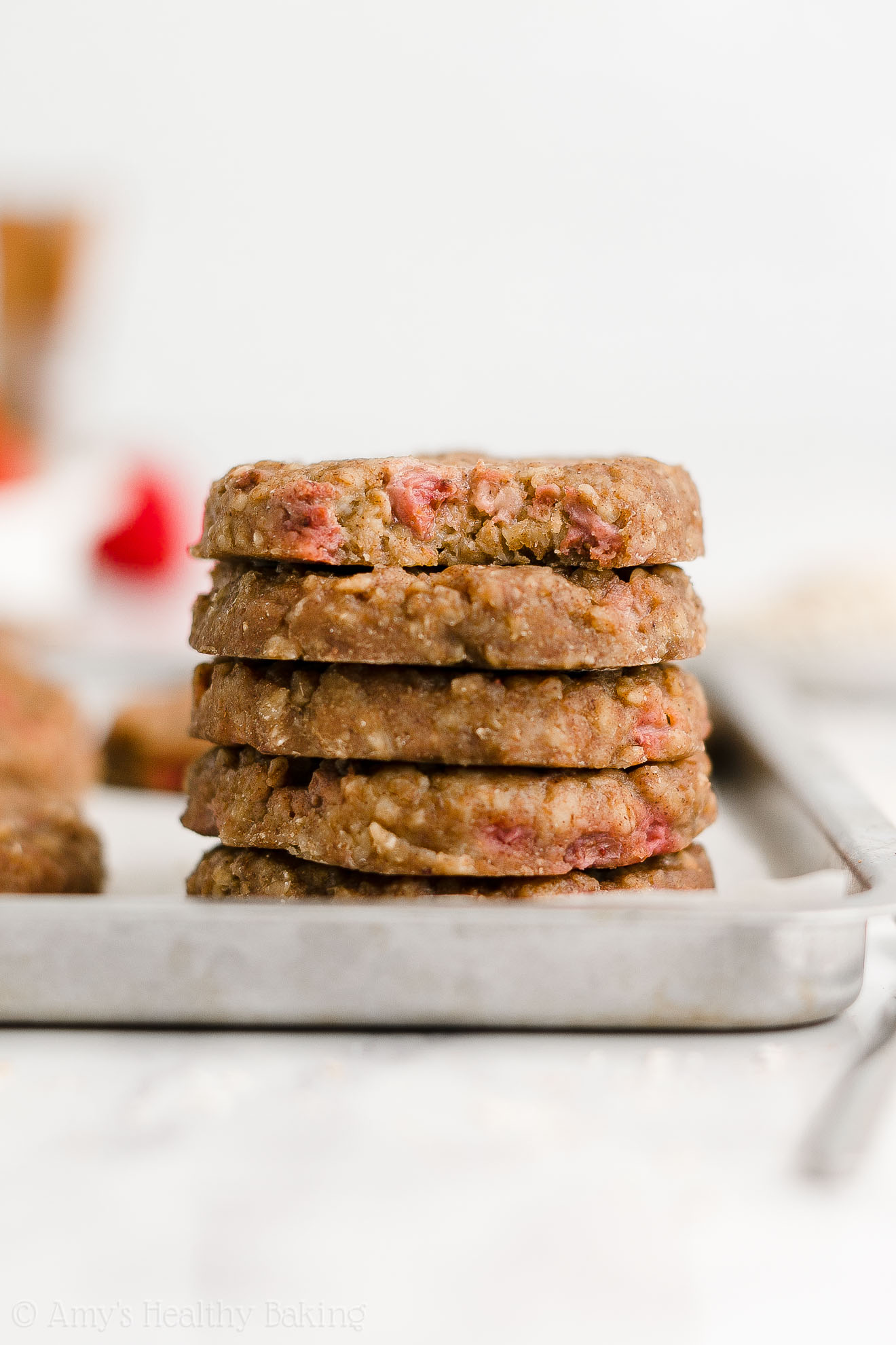 stack of five healthy strawberry oatmeal breakfast cookies on a rimmed baking sheet, with a bite missing from the top cookie to reveal the texture inside – easy strawberry breakfast cookies recipe – soft, chewy, low calorie, low fat, and clean eating with whole wheat, gluten free, dairy free, and nut free options – made with oats, fresh strawberries, pure maple syrup, and no refined sugar