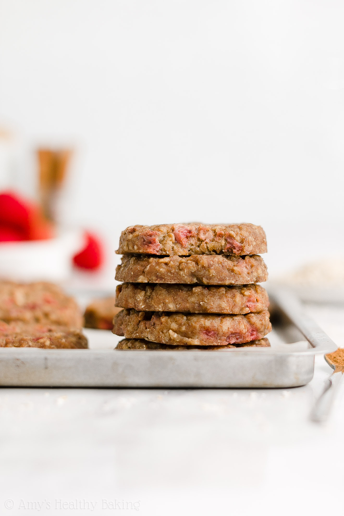 stack of five healthy strawberry oatmeal breakfast cookies inside of a rimmed baking sheet with the top cookie missing a bite to reveal the texture inside, plus a bowl of strawberries and jar of cinnamon sticks in the background – easy strawberry breakfast cookies recipe – soft, chewy, low calorie, low fat, and clean eating with whole wheat, gluten free, dairy free, and nut free options – made with oats, fresh strawberries, pure maple syrup, and no refined sugar