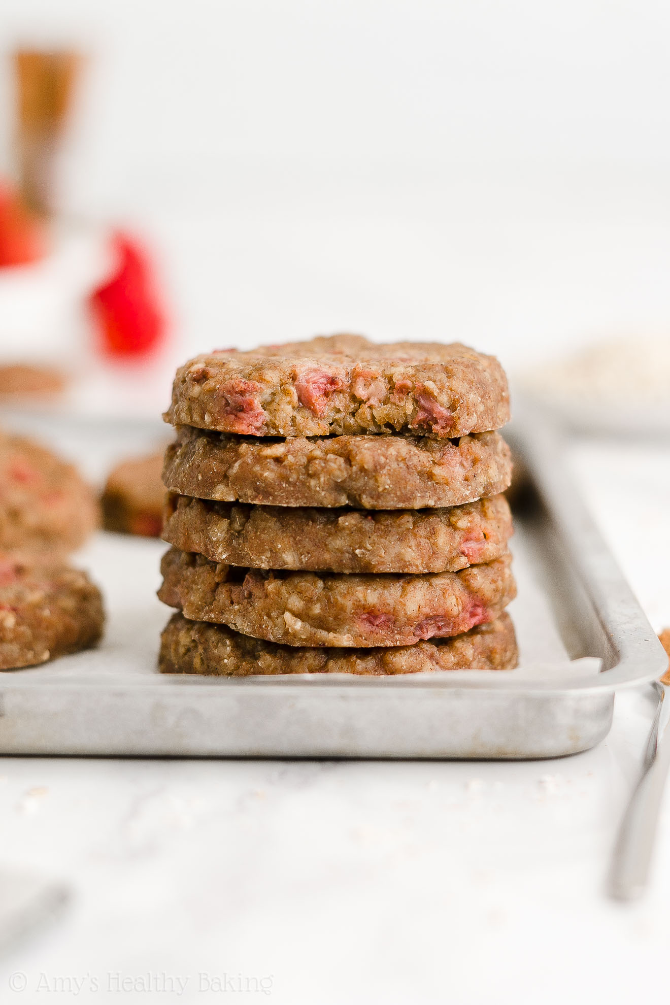 a stack of five healthy strawberry oatmeal breakfast cookies sitting on a rimmed baking sheet, with the top cookie missing a bite to reveal the texture inside – easy strawberry breakfast cookies recipe – soft, chewy, low calorie, low fat, and clean eating with whole wheat, gluten free, dairy free, and nut free options – made with oats, fresh strawberries, pure maple syrup, and no refined sugar