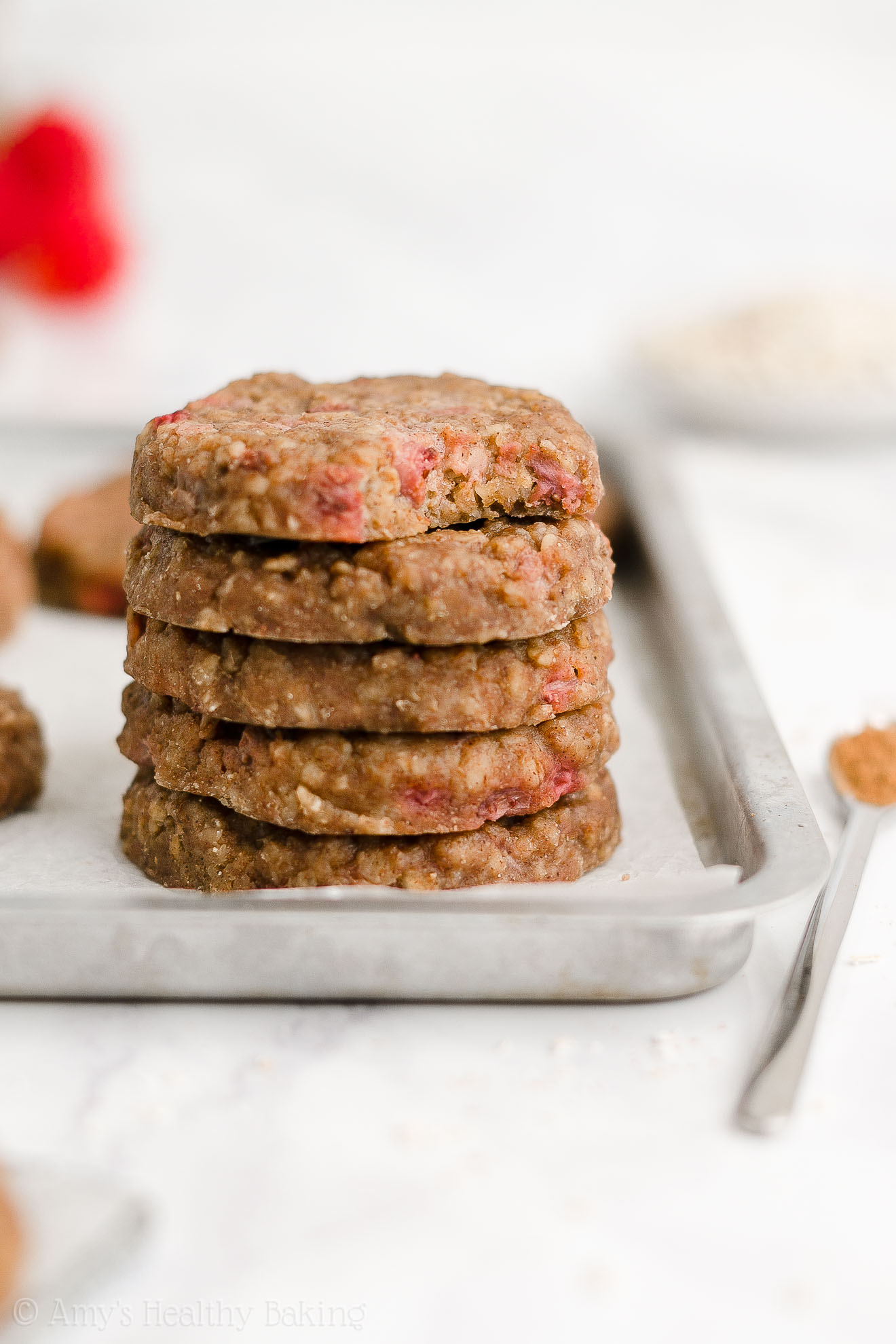 stack of five healthy strawberry oatmeal breakfast cookies sitting in a rimmed baking sheet with the top cookie missing a bite to reveal the texture inside, along with a small bowl of oats and some fresh strawberries in the background – easy strawberry breakfast cookies recipe – soft, chewy, low calorie, low fat, and clean eating with whole wheat, gluten free, dairy free, and nut free options – made with oats, fresh strawberries, pure maple syrup, and no refined sugar